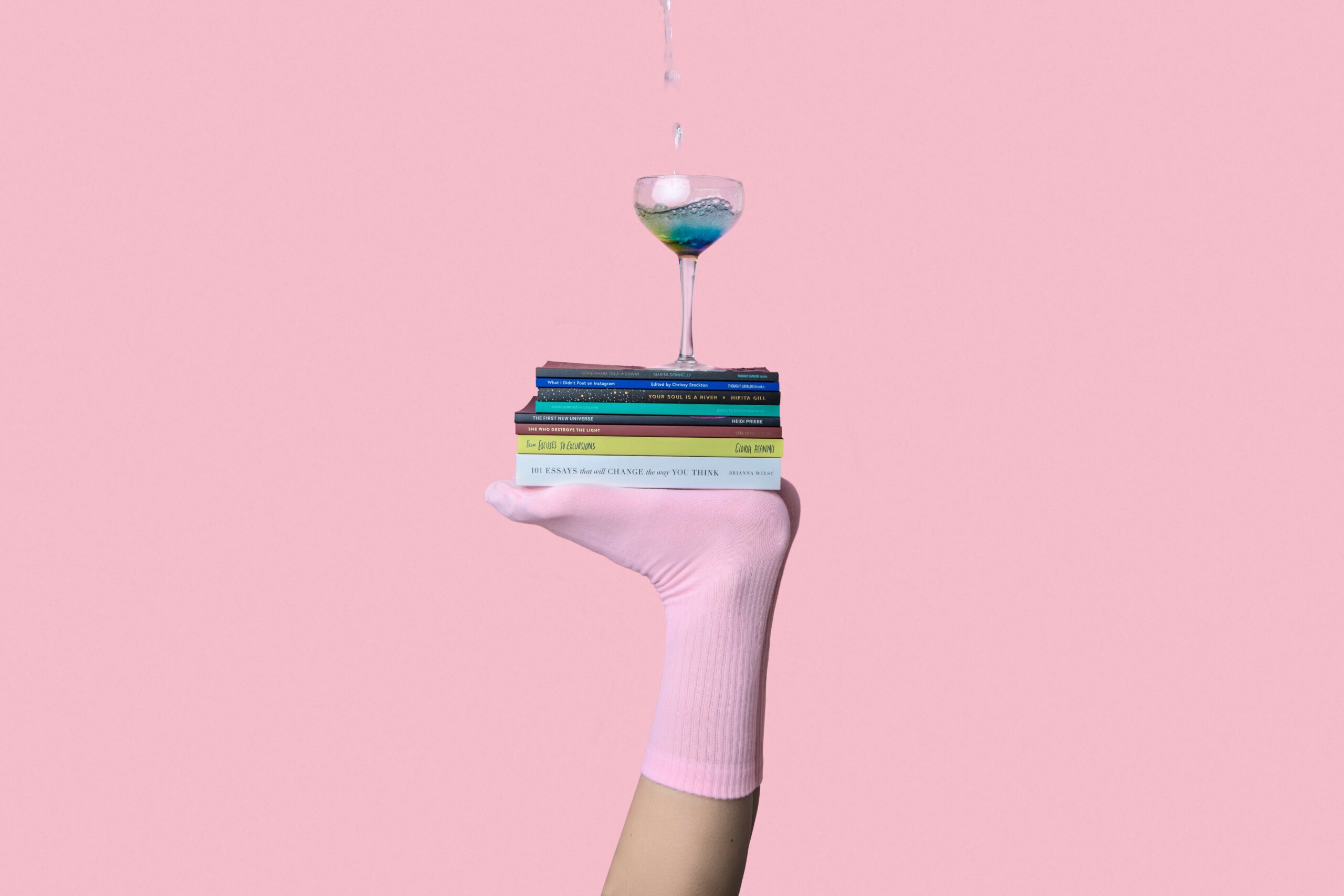 foot with a pink sock holding up books and a martini glass on a pink background