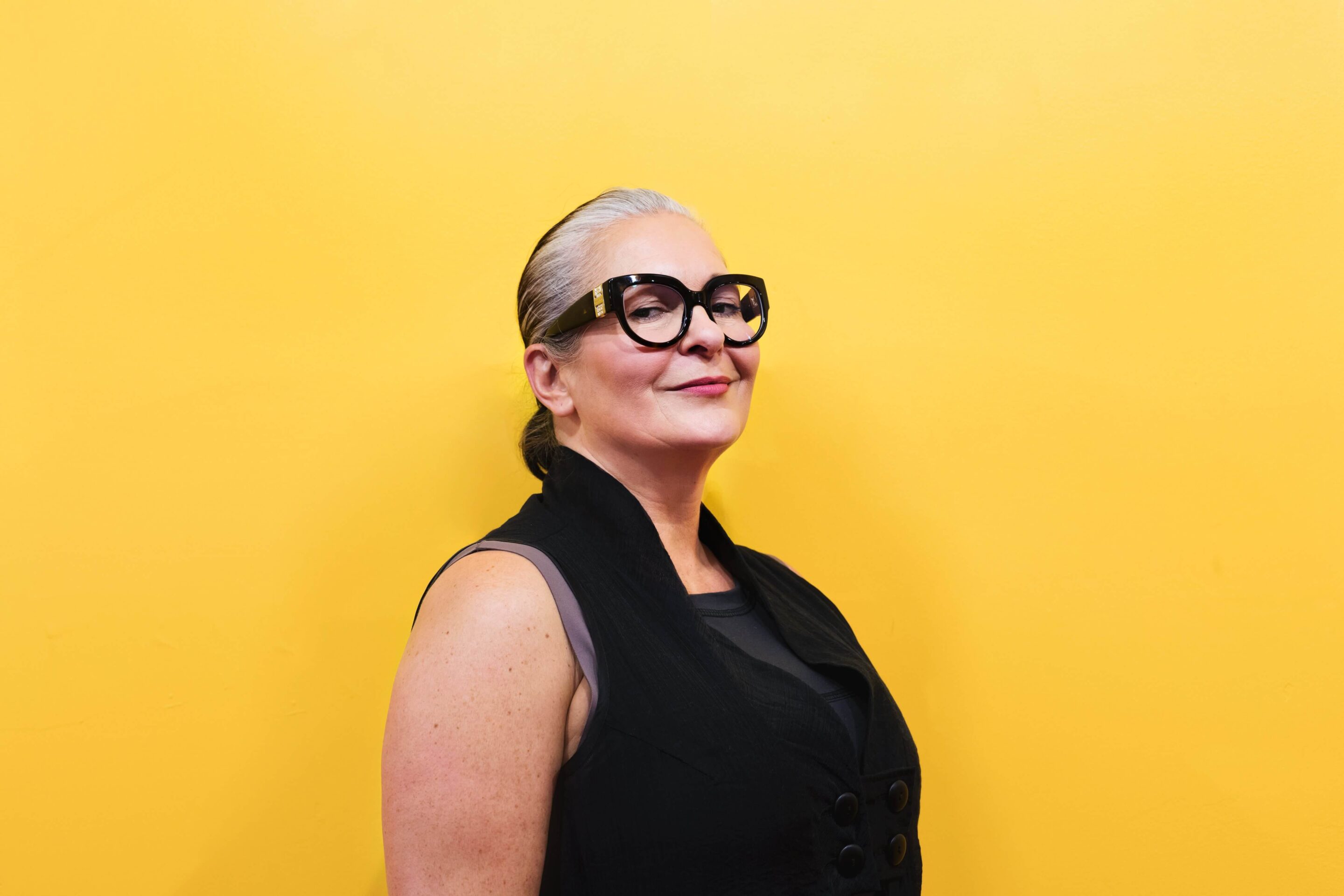 fashionable older woman in glasses stands in front of a yellow background